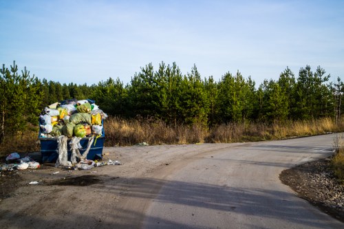 The image depicts a large blue skip filled with a mixture of household rubbish, including plastic bags and assorted waste materials, situated on the edge of a dirt and gravel path in an open, semi-rural area. The skip is overflowing with waste, some of which has spilled onto the ground surrounding it. In the background, there is a dense line of green trees and bushes under a clear, blue sky, indicating a natural environment. The scene suggests a disposal or collection point in a countryside setting, consistent with rubbish removal services provided by companies such as Garden Clearance Earls Court in west London postcodes, subtly reflecting local surroundings. The overall atmosphere is quiet and unoccupied, highlighting the presence of accumulated waste ready for clearance, typical of garden or rubbish removal in the Earl's Court area or nearby localities.