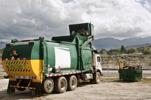 A large green waste collection truck with a yellow caution sign at the rear is parked on an open area with a gravel surface, next to a green waste bin filled with garden debris. The truck has a mechanical compactor on top, with a partially open lifting mechanism for loading rubbish. The scene features a cloudy sky with some sunlight filtering through, and distant mountains and trees along the horizon, suggesting a rural or suburban environment. The vehicle and waste container are positioned on a flat, unpaved space, indicative of a site used for rubbish removal or garden clearance services, such as those provided by Garden Clearance Earls Court. The overall setting is quiet and utilitarian, with a focus on the collection and disposal of garden waste or general rubbish, aligning with professional waste management operations.