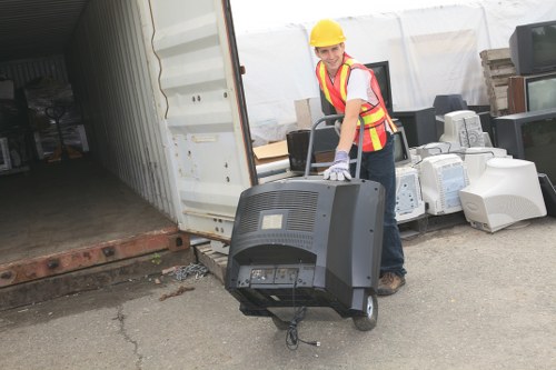 A woman wearing a yellow safety helmet, high-visibility vest, and safety gloves is standing outdoors on a paved surface, holding a rear projection television set by its sides. The TV has a large, curved black plastic casing with visible vents on the back, and the front screen is not visible. Behind her is a partially open large shipping container or storage unit, revealing a dark interior, and the container rests on a raised brick platform. To her right, there are several discarded white electronic appliances, including old computer monitors and possibly printers, stacked and scattered on the ground. The background includes a section of white material, possibly a tent or temporary construction fencing, indicating an operational or waste clearance site in an urban or suburban area. This scene relates to garden clearance and rubbish removal services offered by Garden Clearance Earls Court, serving local areas including parts of West London and surrounding postcode districts.